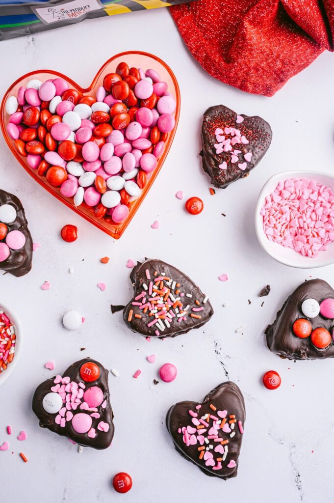 vertical image of chocolate covered apple hearts on counter with m&ms and sprinkles