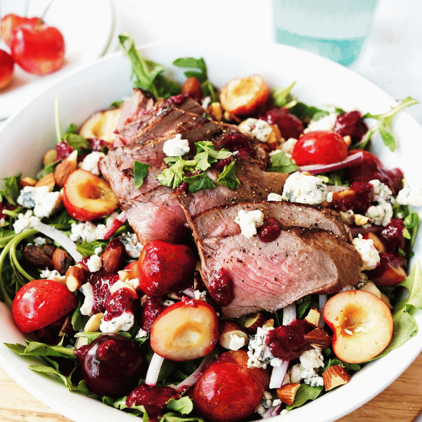 Steak & Arugula Salad with Cherries and Almonds in a white bowl surrounded by cherries