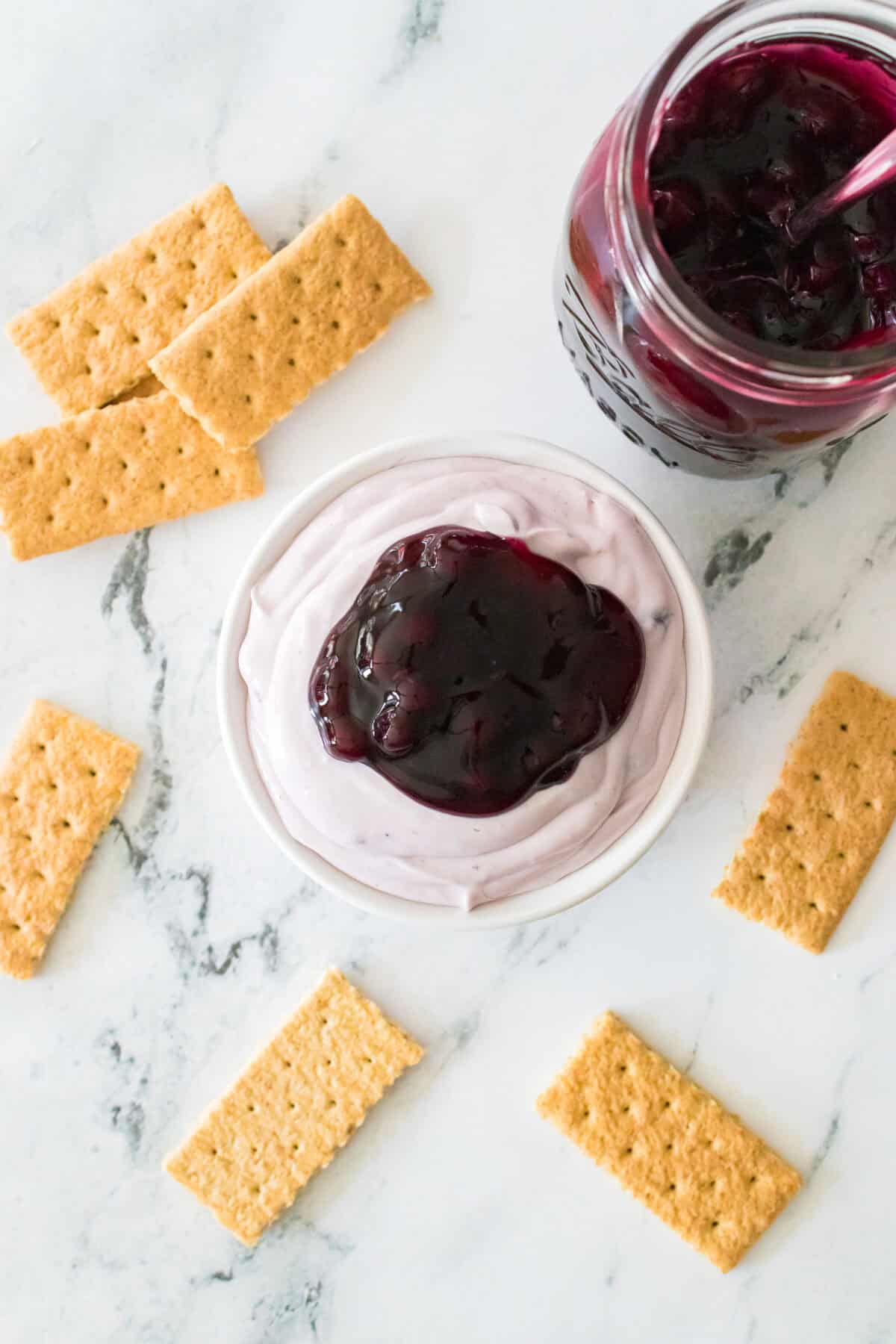 blueberry cheesecake dip in serving dish with graham crackers scattered around for serving with mason jar of blueberry pie filling