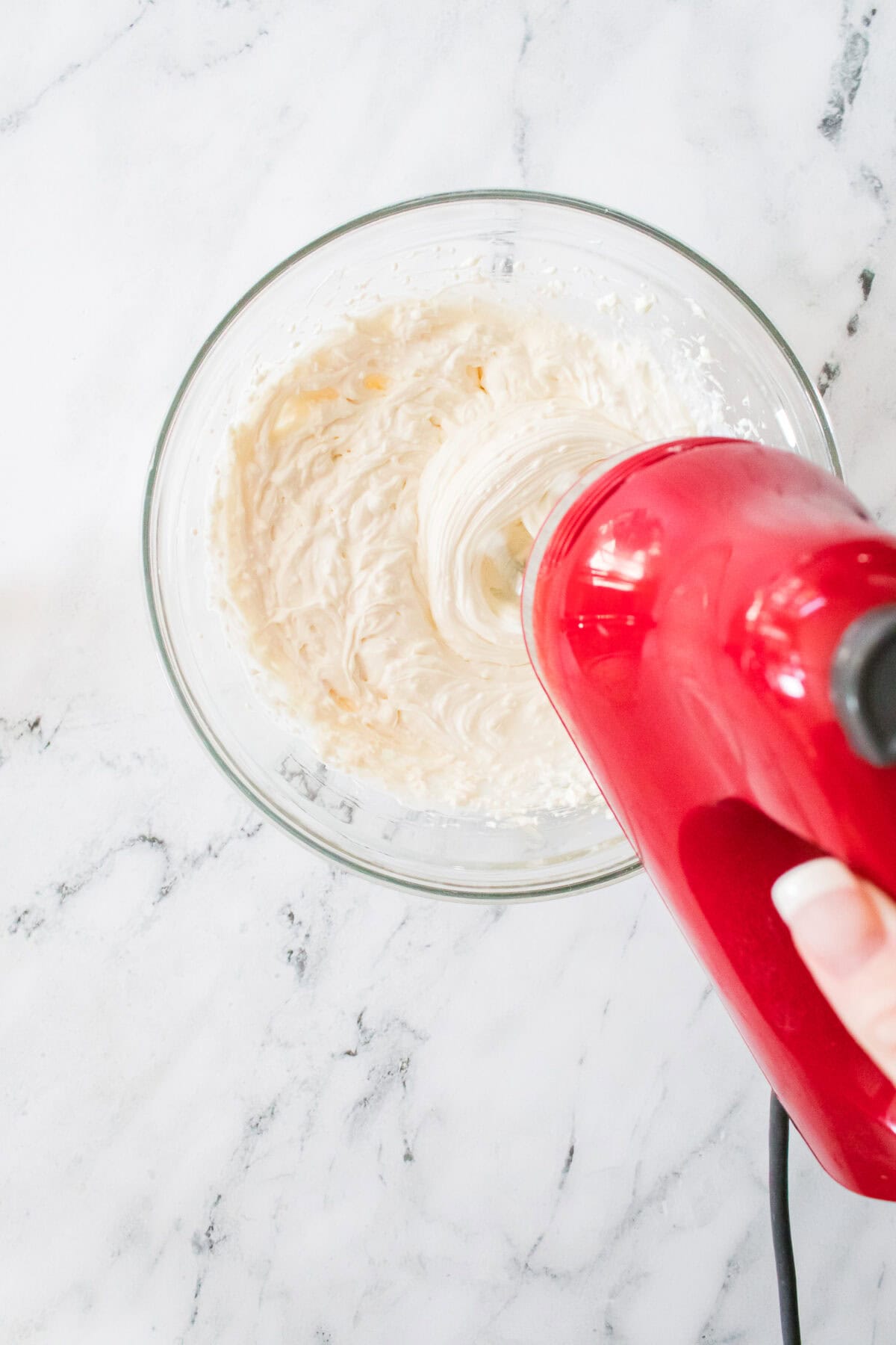 handheld mixer mixing together cream cheese, greek yogurt and vanilla extract in glass bowl