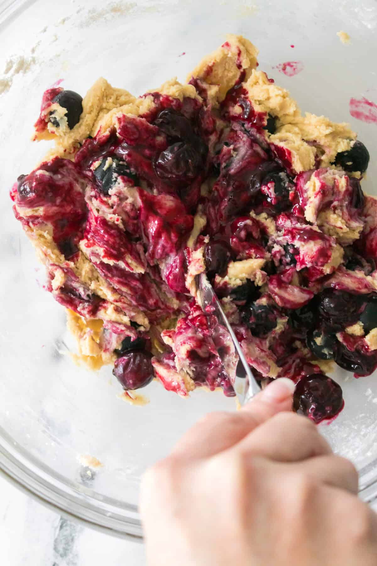 close up vertical shot of hand holding butter knife and blending blueberry jam into cookie dough with fresh blueberries